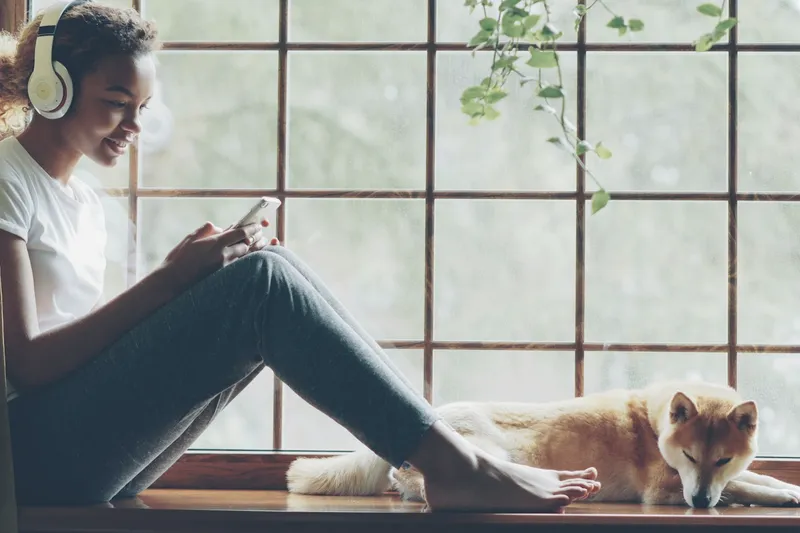 A woman with headphones sits on a window sill looking at her phone, a dog asleep beside her.